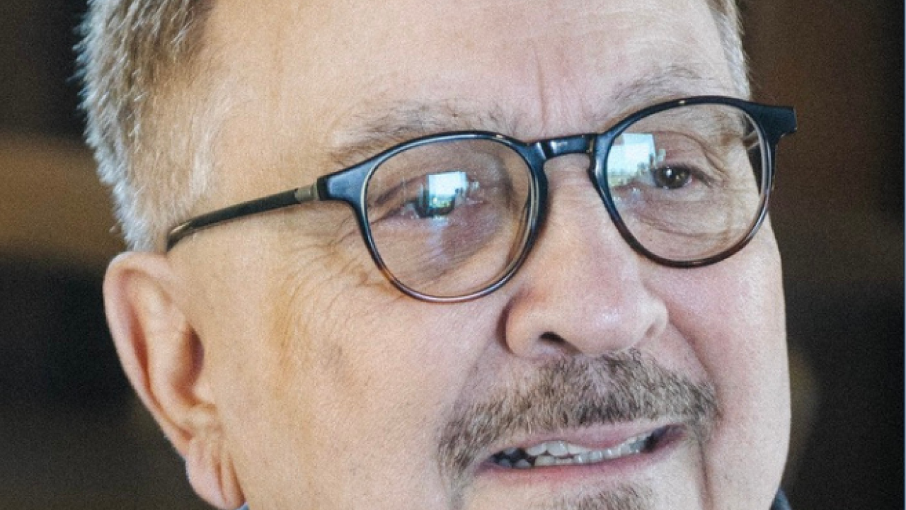 A smiling older man with glasses, wearing a dark sweater, against a blurred background.