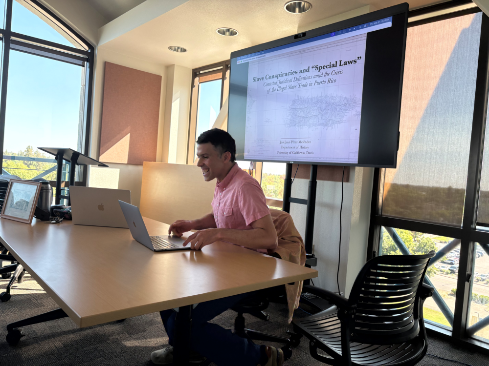 Man in pink shirt at conference table with laptop and projected presentation