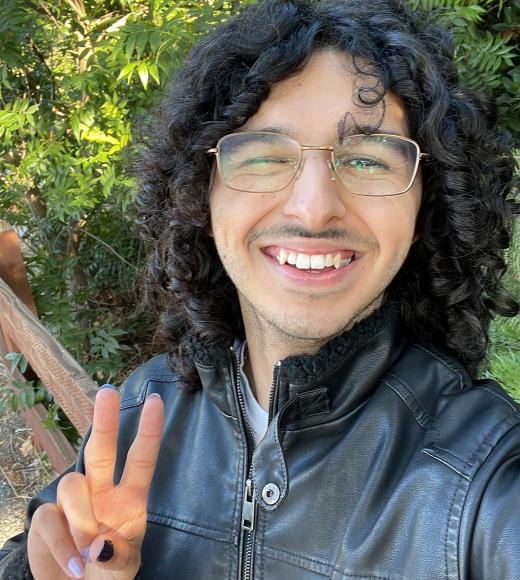 Smiling person with curly hair and glasses, flashing peace sign, leather jacket, leafy background
